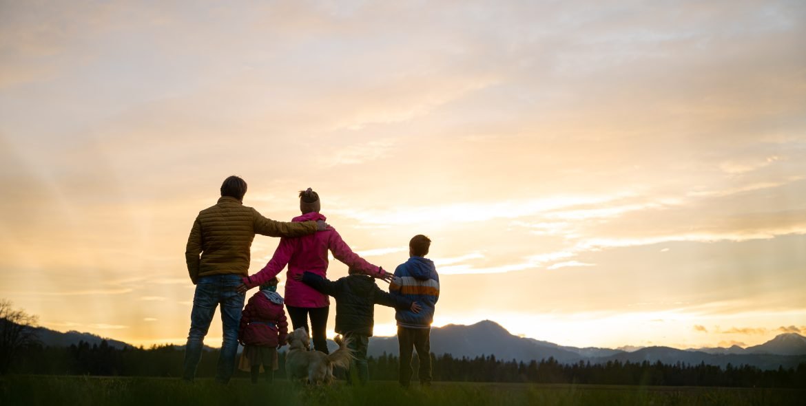 View from behind of family of five and a dog standing outside at dusk with teir arms around each other.