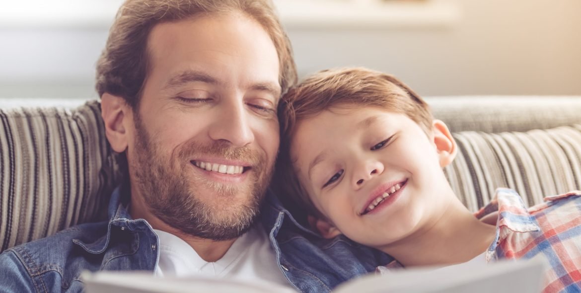 Father and son are reading a book and smiling while spending time together at home
