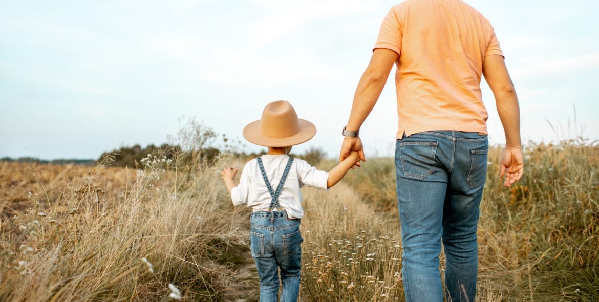 Father and young son walking keeping hands together on the field during the summer activity, back view