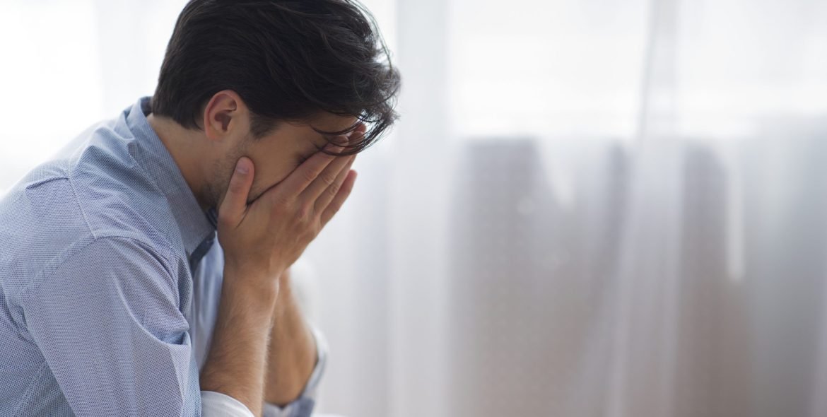 Male Depression. Unrecognizable Frustrated Man Covering Face With Hands Crying Sitting Against Window. Copy Space