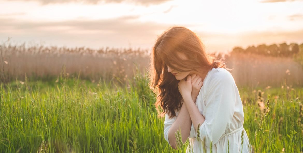 Girl closed her eyes, praying in a field during beautiful sunset. Hands folded in prayer concept for faith, spirituality and religion. Peace, hope, dreams concept