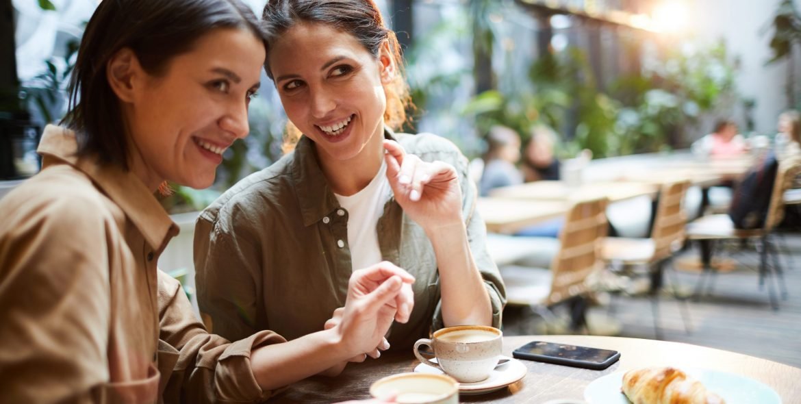 Cheerful young woman in casual shirts sitting at table and drinking coffee while gossiping about people in cafe