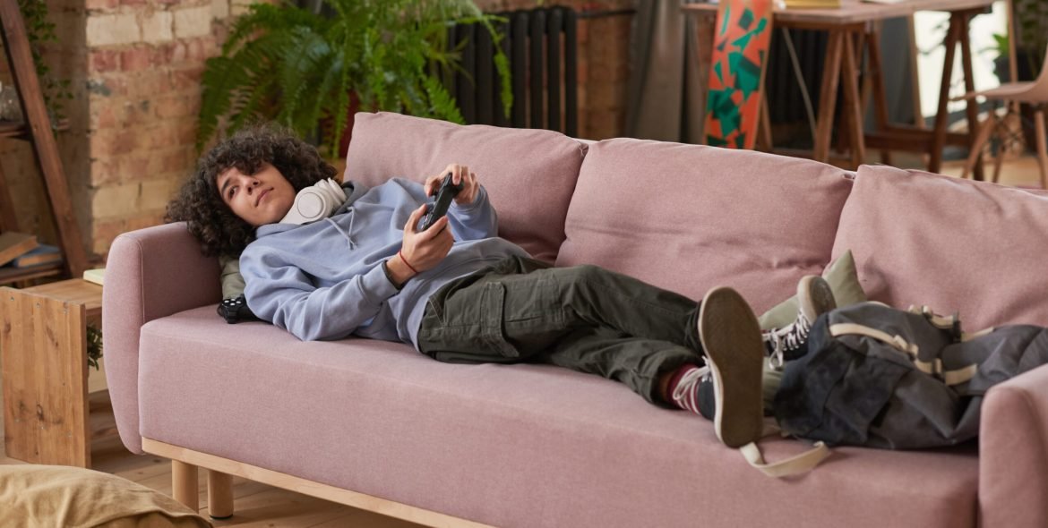 Guy lying on the sofa with joystick and playing video game during his leisure time in the room