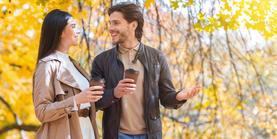 Happily married young couple with cups of coffee walking by autumn forest, having conversation, copy space