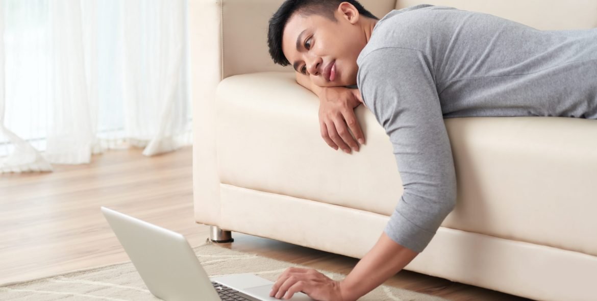 Lazy yuong Vietnamese man lying on sofa and watching something on laptop