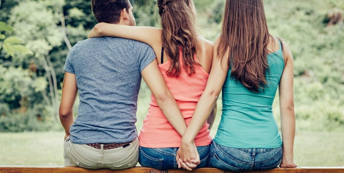 Love triangle, a girl is hugging a guy and he is holding hands with another girl, they are sitting together on a bench