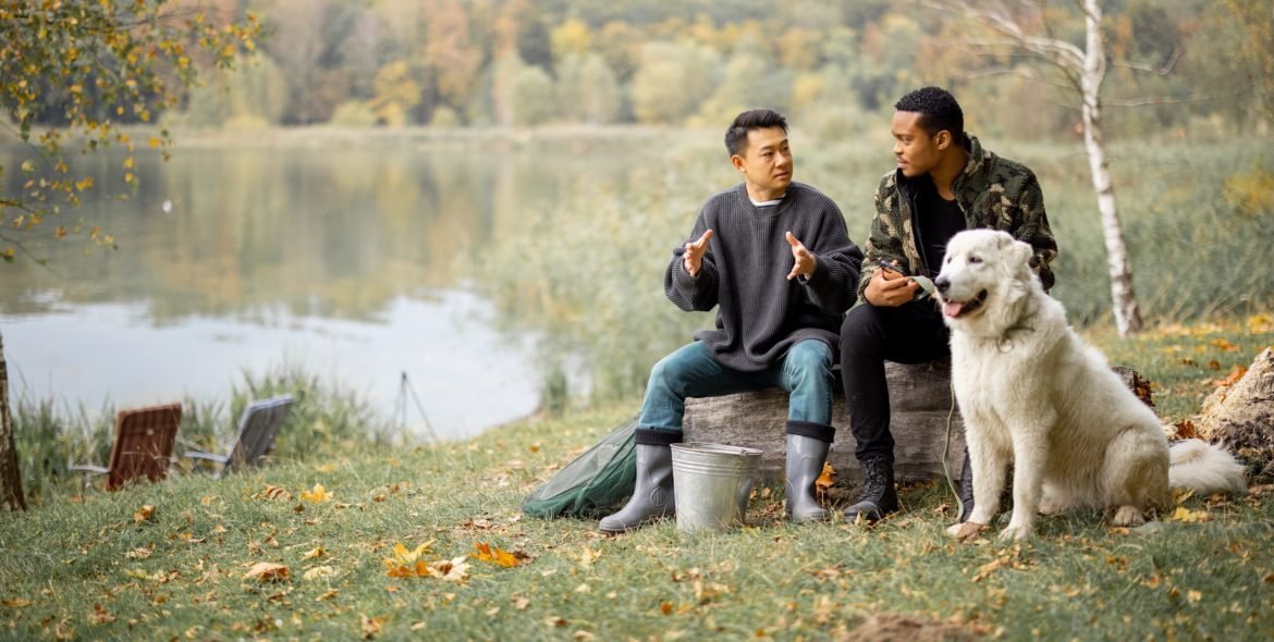 Multiracial male friends talking while sitting on fallen tree near Maremmano-Abruzzese Sheepdog on lake or river coast. Concept of rest and weekend in nature. Idea of friendship. Autumn day