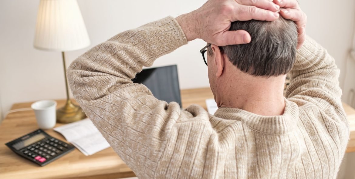 Senior man worried about bills sitting at home holding hands on head being stressed with debts.