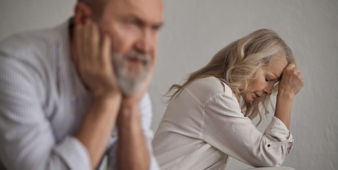 Frustrated blonde lady and a silent gray-haired male sitting apart from each other on the bed