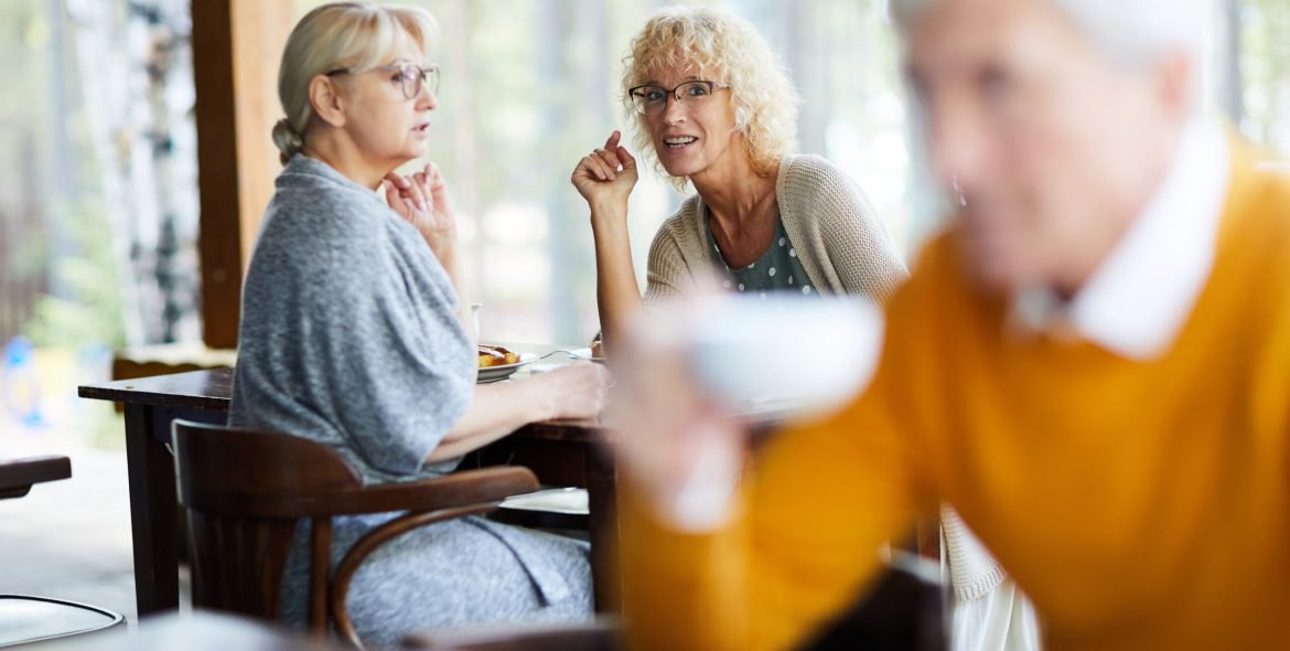 Content attractive mature ladies in casual clothing sitting at table and gossiping about handsome man in cafe, curly-haired lady pointing at this man