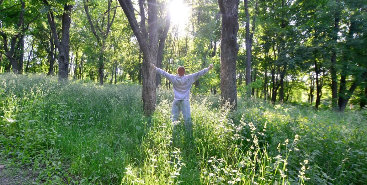 A young guy in a gray sports suit rejoices in the rising of the sun among the trees in the forest. Recreation during a sports run in the open air forest. The delight of a beautiful dawn