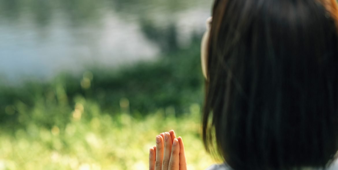 Mindful Young woman, feeling grateful, meditating, sitting by the water, hands in prayer position