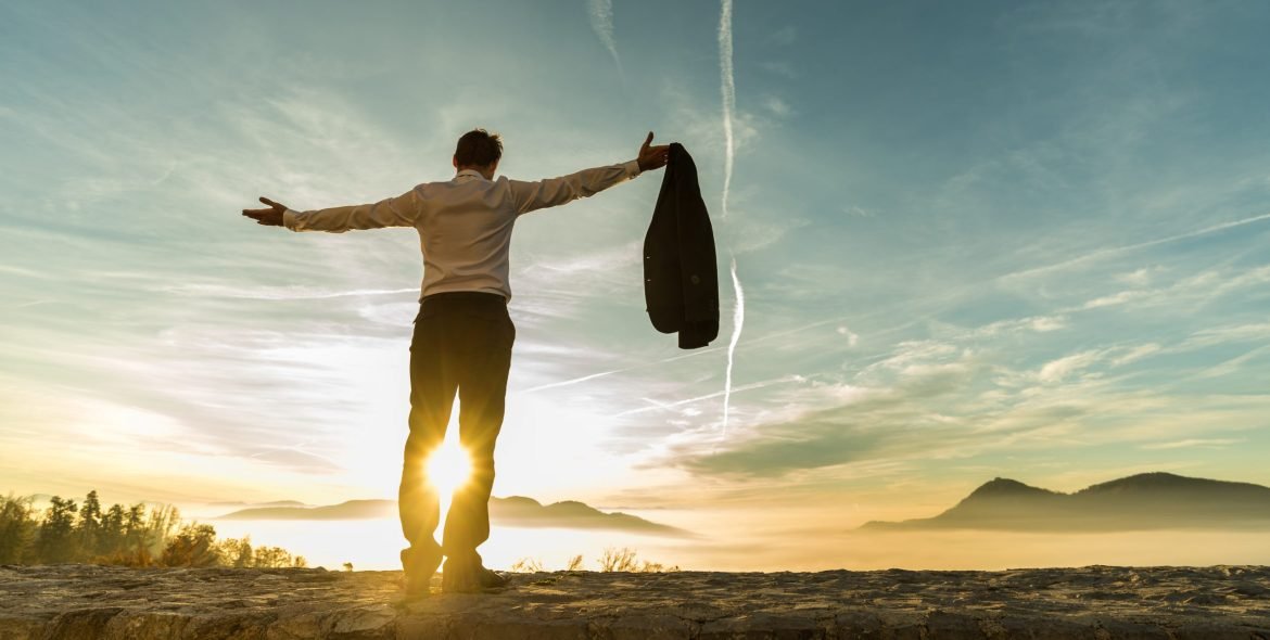Rear view of successful businessman rejoicing at sunrise standing against the bright glow of the sun with outstretched arms.