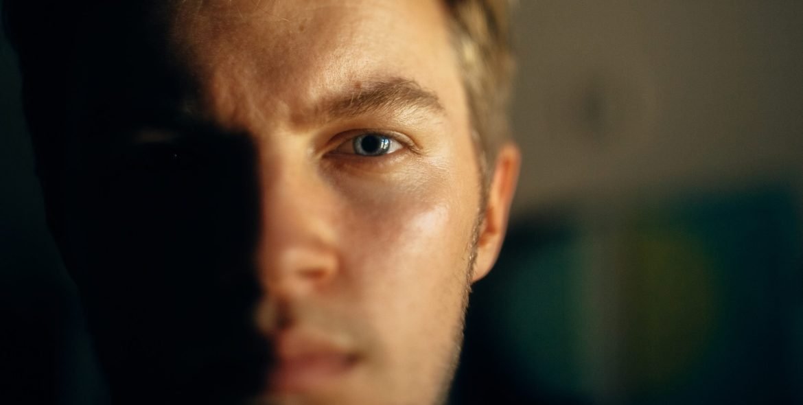 Serious man portrait in light on dark background. Closeup of man face with blue eyes and blonde hair in softlight. Selective focus. Creative image