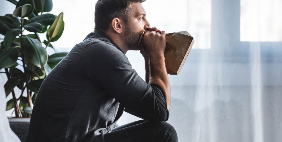 side view of handsome man with panic attack breathing in paper bag in apartment