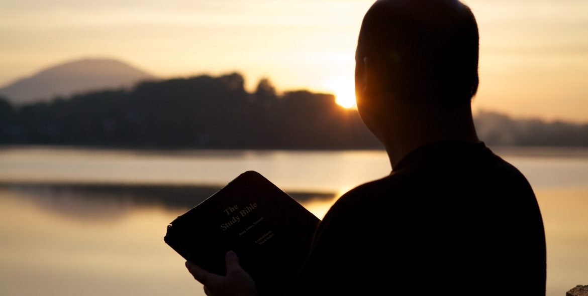 A silhouette of a man holding the bible surrounded by hills and the sea during the sunset