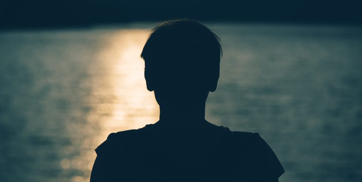 Silhouette of depressed sad woman standing by the lake in cold autumn sunset. Gloomy adult female person looking at water. Loneliness and solitude concept.