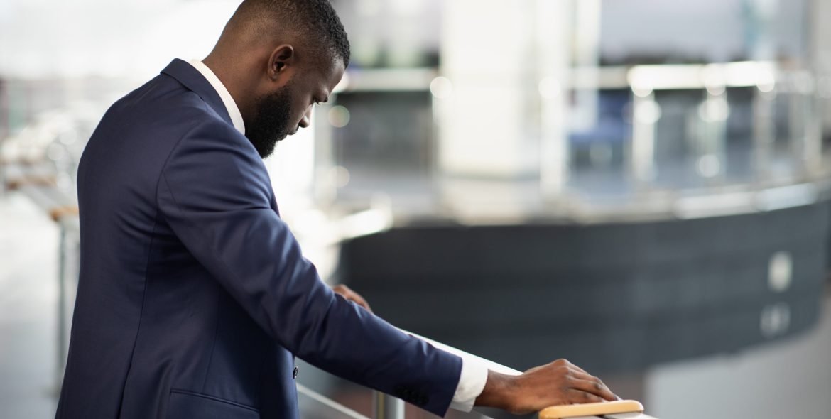 Stressed african american young man in suit entrepreneur standing by railing with head down, experiencing difficulties at work, back view shot, panorama with copy space, financial crisis and business