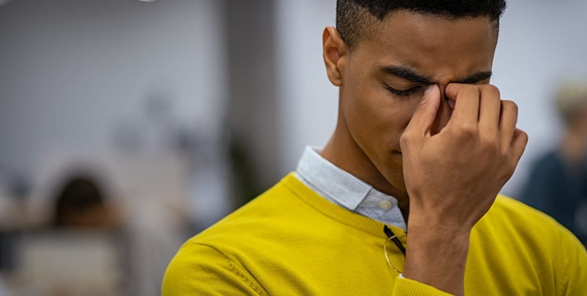 Portrait of an upset young businessman standing in creative office. Black depressed business man with a terrible headache. Stressed african man feeling strain in eyes after working for long hours on computer: overwork, burnout and workload concept.