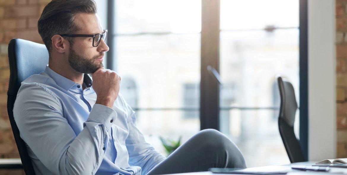 Calm handsome Caucasian man looking away while sitting in the office at daytime and touching his chin