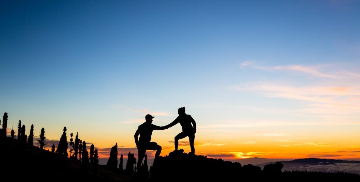 Teamwork couple with helping hand and trust. Silhouette of team help over mountains sunset. Man and woman hikers or climbers reaching goals in inspirational landscape on Tenerife Canary Islands.