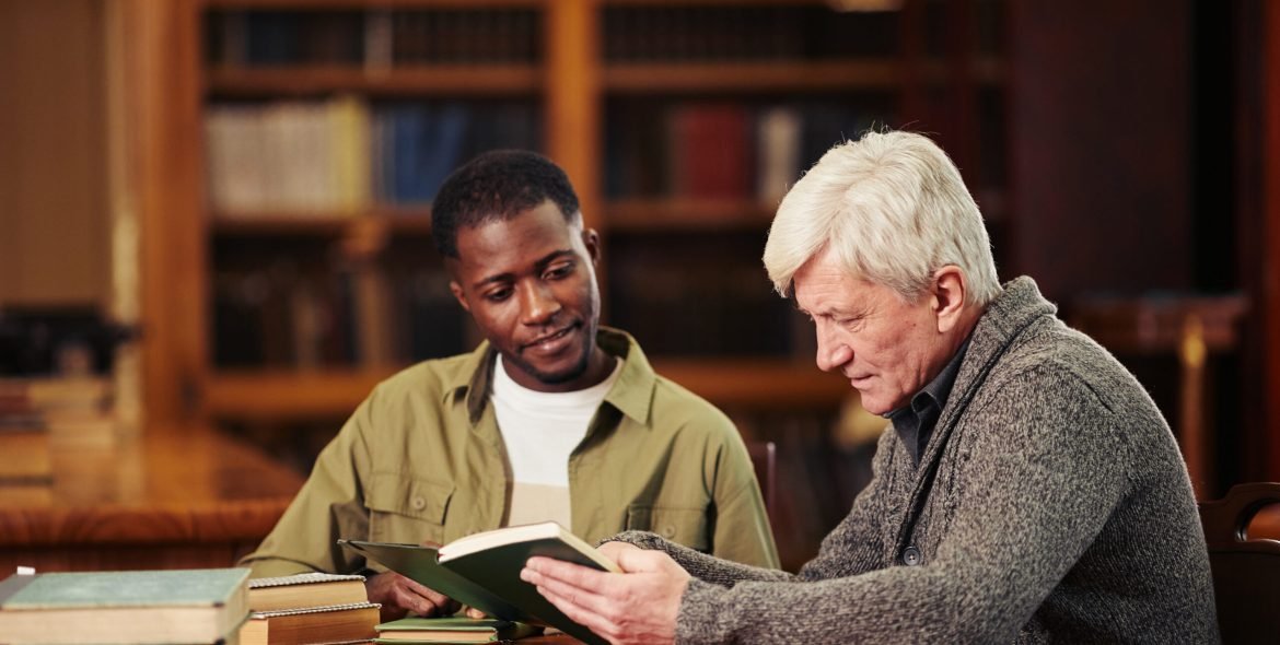 Portrait of male senior student reading book in library while preparing for exam with African-American friend