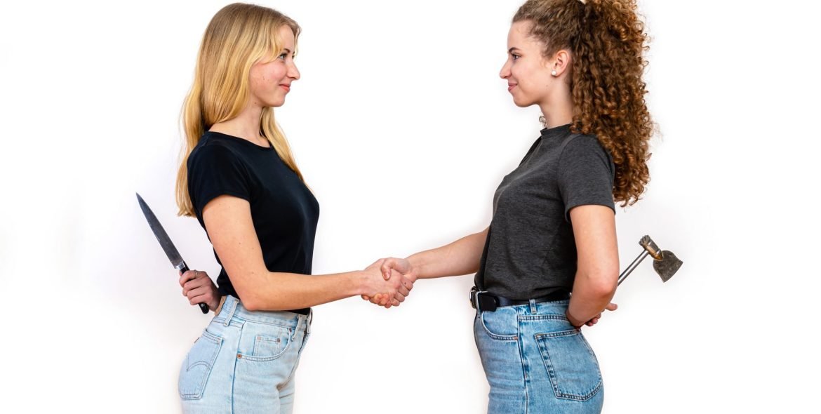 two smiling women standing face to face and hiding knife and axe behind back, isolated on white background, the concept of hidden hatred and hypocrisy