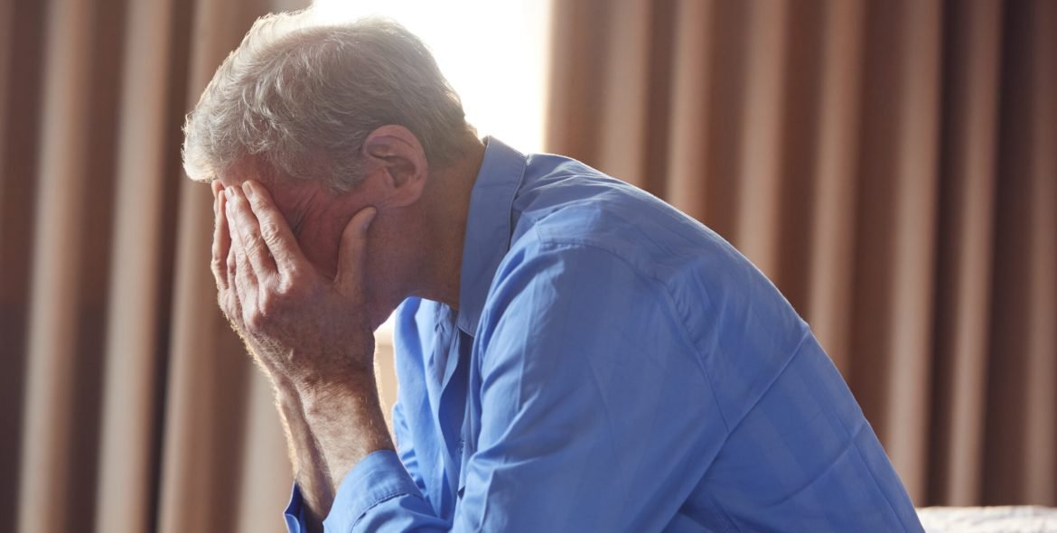 Unhappy And Depressed Senior Man With Head In Hands Sitting On Edge Of Bed At Home