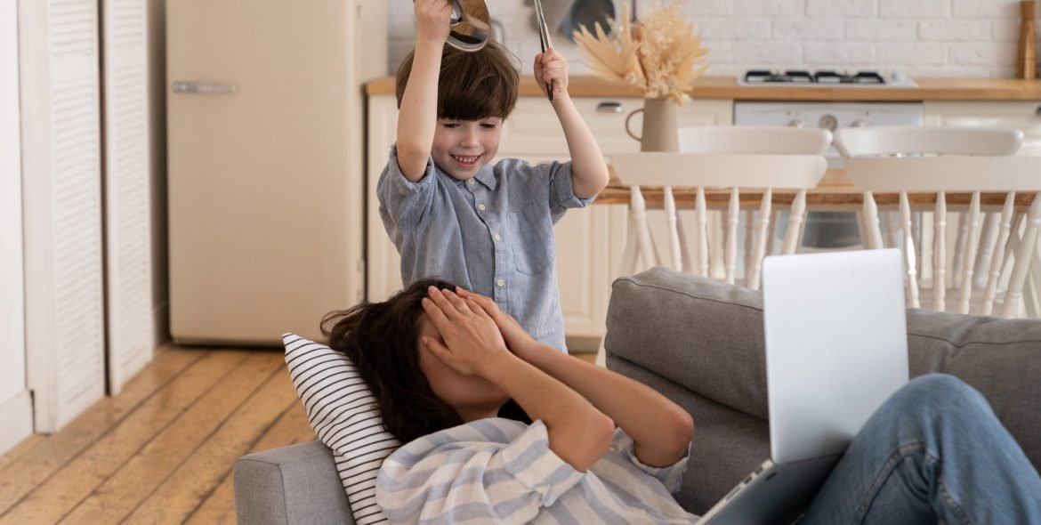 Depressed unhappy mom cover face with hand to protect from noisy son bad behavior. Young mother entrepreneur or freelancer tired of work from home with little kid. Desperate mommy suffer from headache