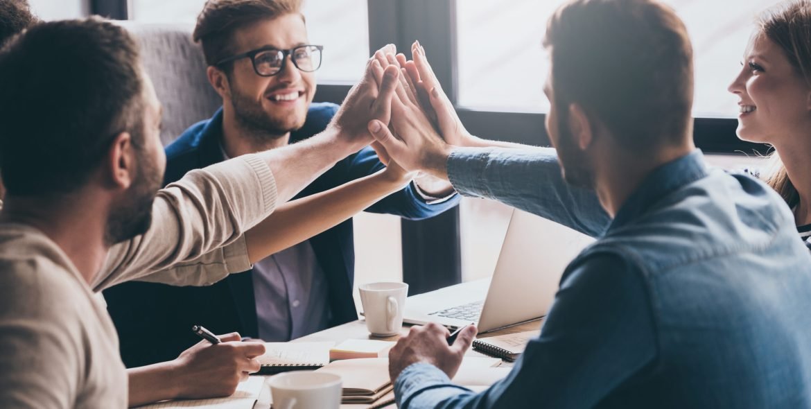 We are leaders on the market! Cheerful young people giving each other high-five with smile while sitting at the office table on business meeting