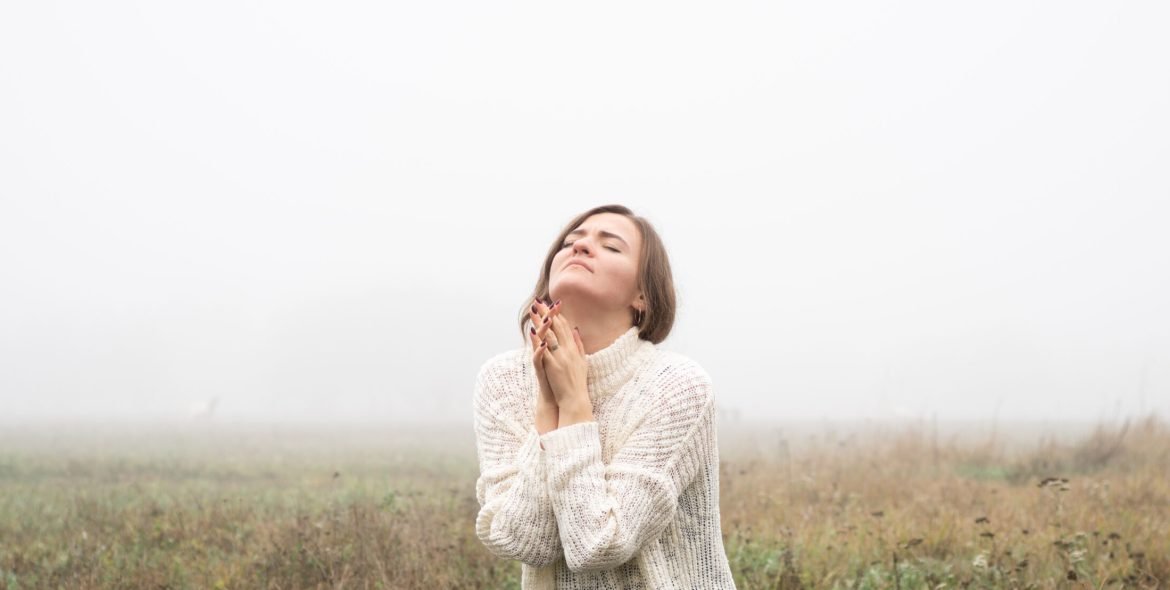 Girl closed her eyes, praying in a field during beautiful fog. Hands folded in prayer concept for faith, spirituality and religion. Peace, hope, dreams concept