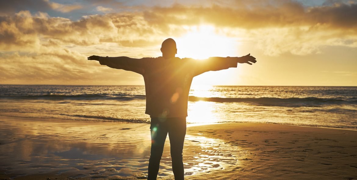 Worship, sunrise and silhouette of man at the beach standing with arms raised. Faith, religious and.