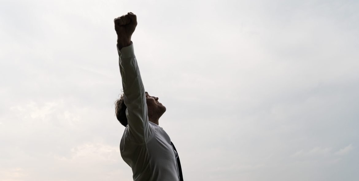 Profile view of successful young businessman standing under cloudy grey sky celebrating success with his arms raised high.