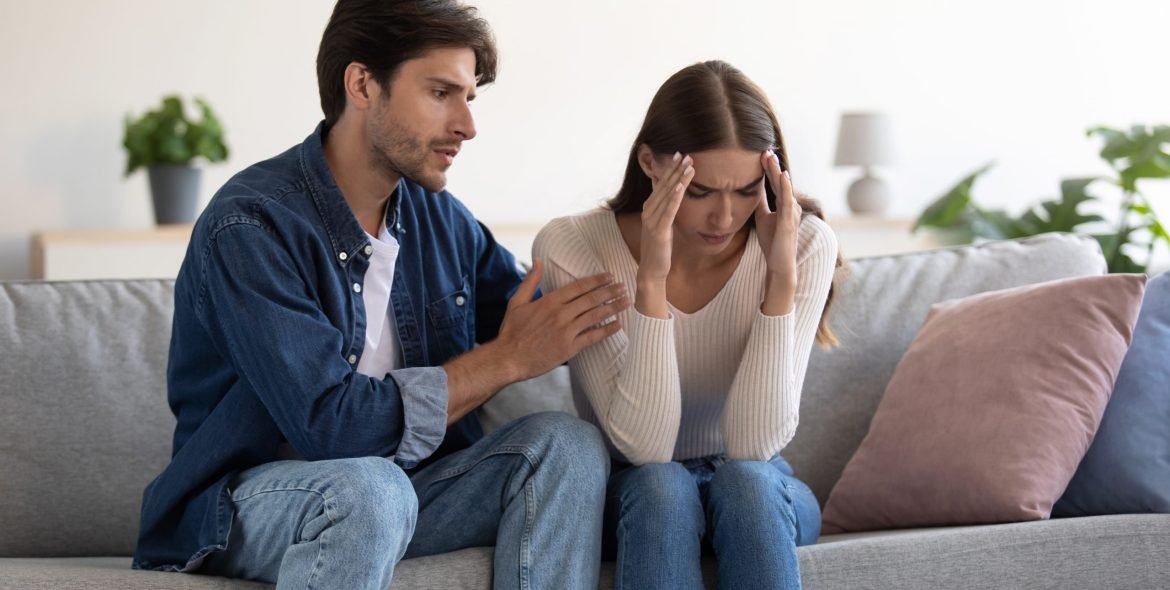 Young caucasian couple sit on sofa, husband calms upset wife at home interior. Loving man caresses lady, caring guy will conquer and make peace with his lover, show support, family overcome problems