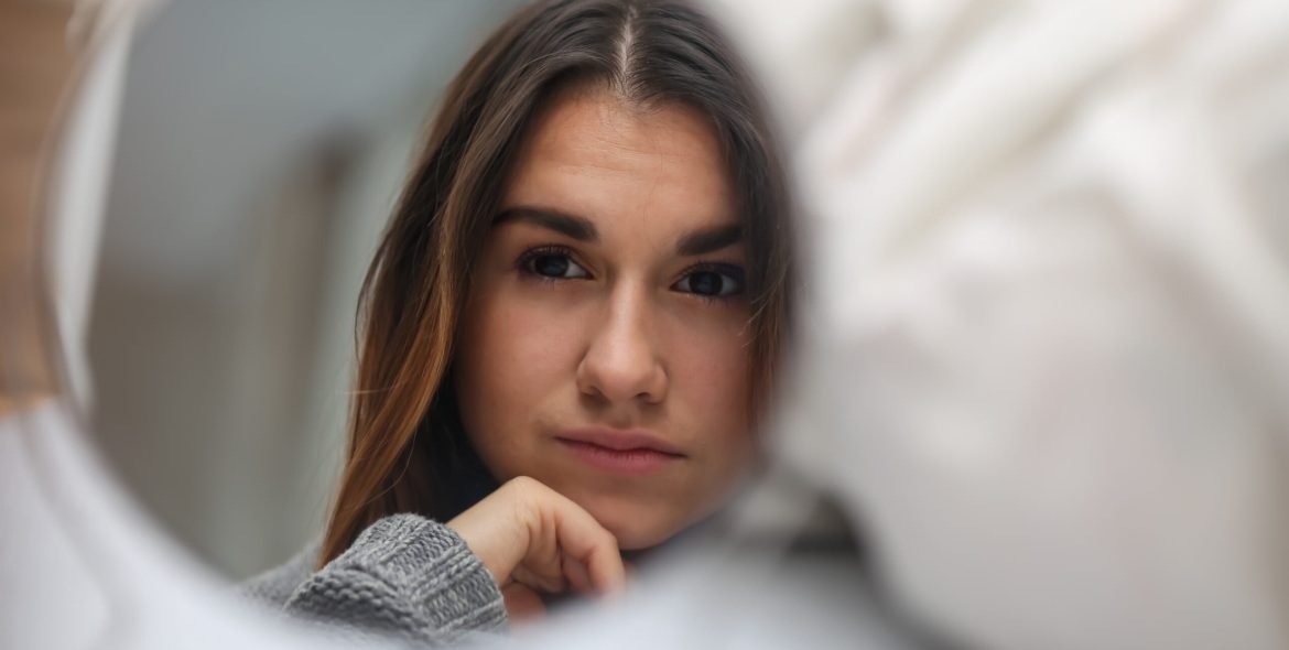 young beautiful girl looking in the mirror, a reflection of the different emotions,white bed in the background