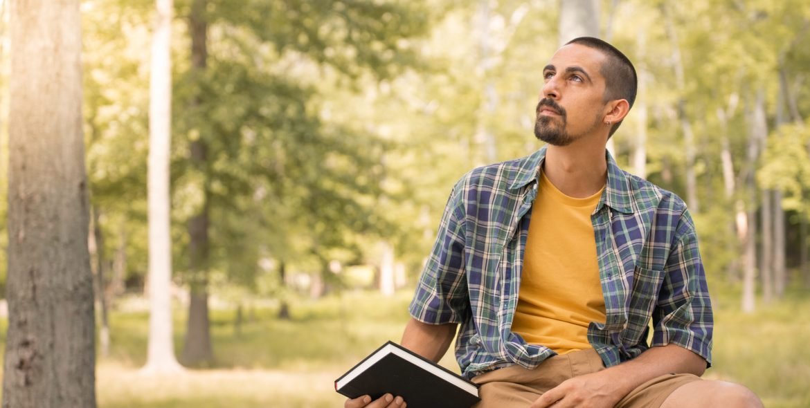 Young man sitting outdoors holding book in a park with trees background - Knowledge faith concept. High quality photo