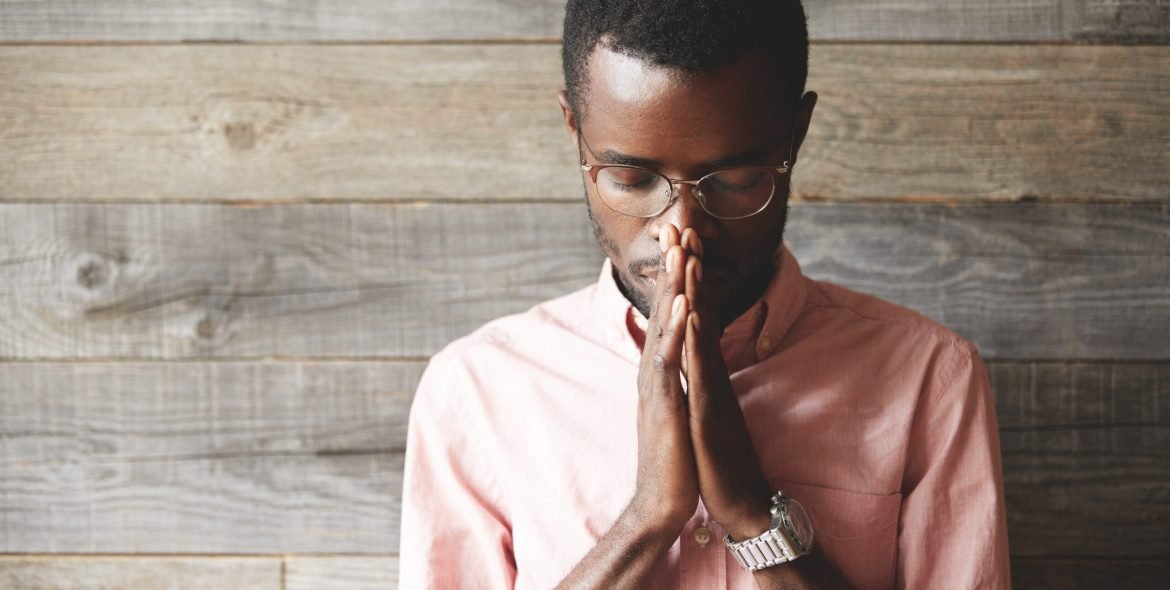 Young meditating and praying African American man wearing pink shirt and glasses, holding hands in prayer against his lips, hoping for the best, asking for wisdom and strenght, with closed eyes