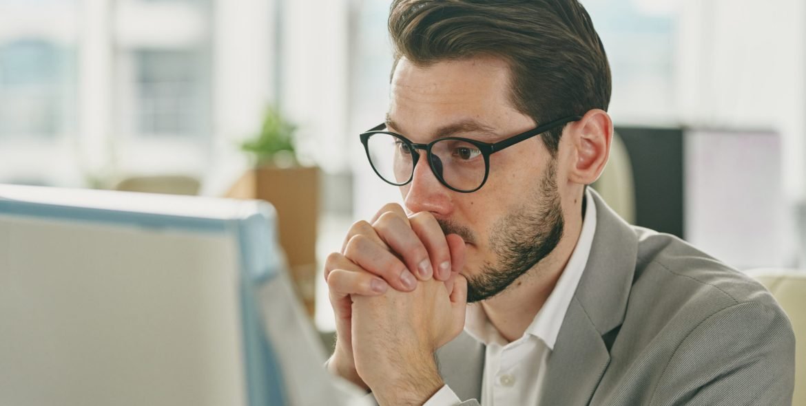 Young pensive businessman in eyeglasses cannot understand information on computer screen while sitting by workplace in office